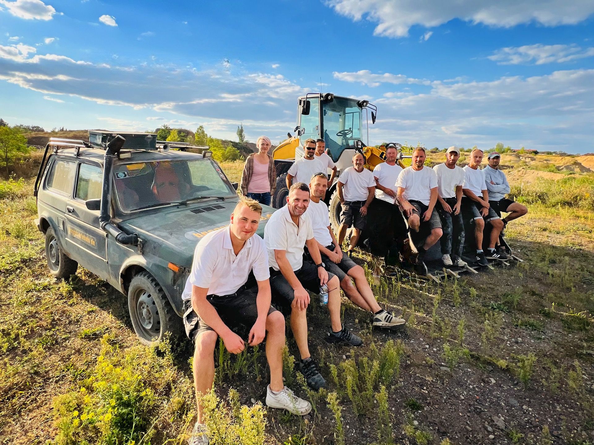 Das Team von Gebr. Rogat in weißen T-Shirts sitzt im Sonnenschein auf einer Wiese vor einem Radlader und einem Geländewagen